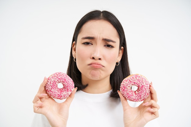 Бесплатное фото Крупный план портрета грустной азиатской женщины, расстроенной диетой, показывающей два соблазненных глазированных розовых пончика