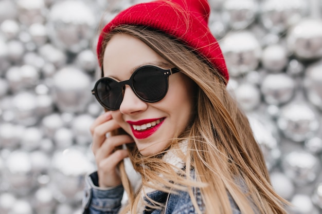 Бесплатное фото Портрет крупным планом улыбающейся удивительной девушки носит черные очки. прекрасная молодая дама в красной шляпе позирует возле дискотечных шаров и трогает ее волосы.