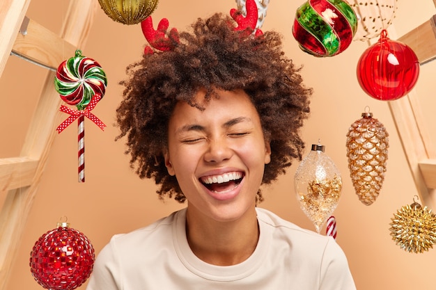 Free photo close up portrait of overemotive curly haired woman with broad smile shows white teeth wears red reindeer horns dressed casually