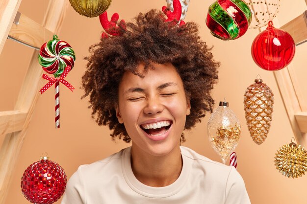 Close up portrait of overemotive curly haired woman with broad smile shows white teeth wears red reindeer horns dressed casually 