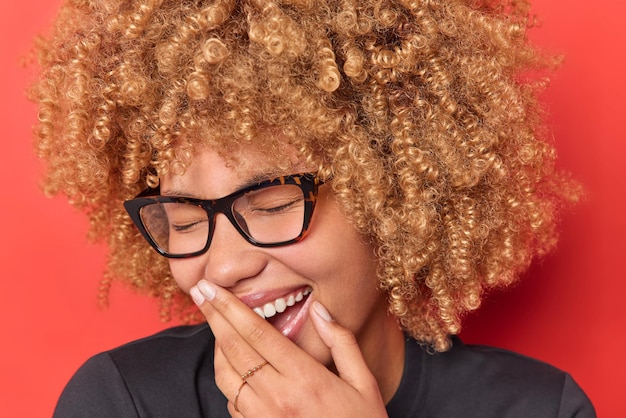 Close up portrait of overjoyed curly haired woman laughs out happily covers mouth with hand keeps eyes closed wears spectacles expresses positive emotions isolated over vivid red background.