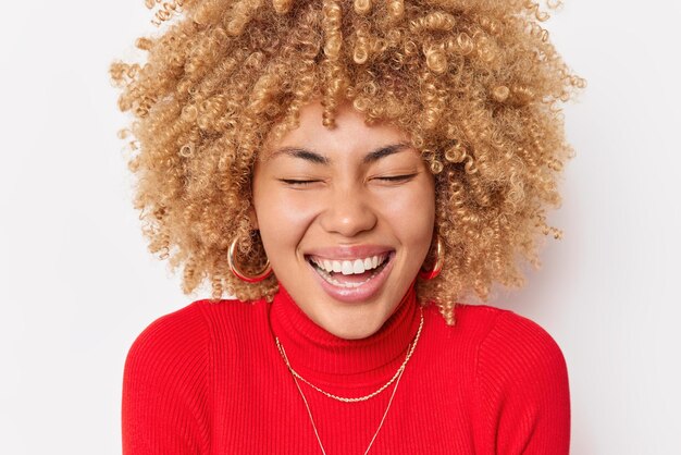 巻き毛の笑いで大喜びの女性の肖像画をクローズアップ幸せにポジティブな本物の感情を表現する白い背景の上に分離された赤いタートルネックとイヤリングを身に着けている幸せと喜び
