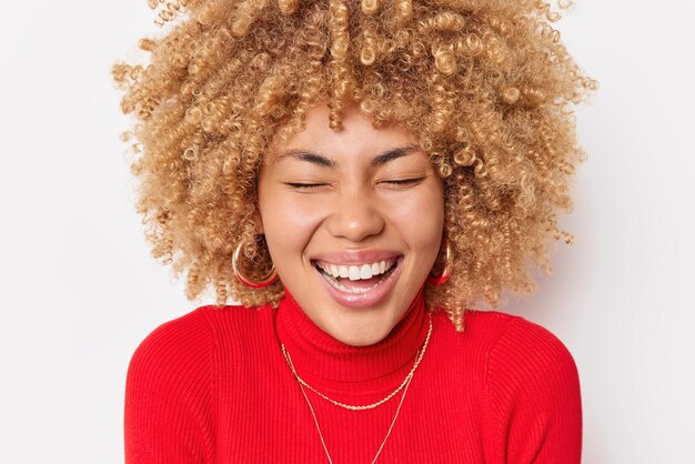 Close up portrait of overjoyed woman with curly hair laughs happpily expresses positive authentic emotions wears red turtleneck and earrings isolated over white background Happiness and joy