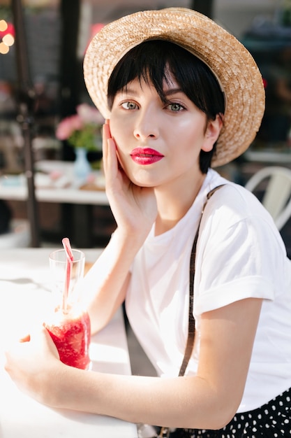 Close-up portrait of pensive girl with gray eyes and red lips resting in cafe with glass of icy beverage