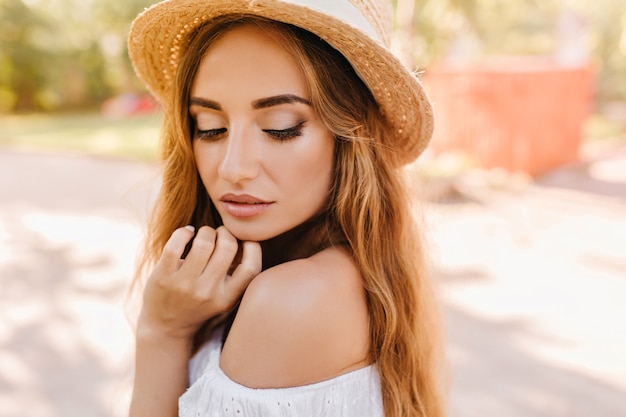 Foto gratuita ritratto del primo piano di donna pensosa con pelle leggermente abbronzata e trucco nudo alla moda in posa con gli occhi chiusi. foto all'aperto della signora con cappello vintage che tocca il mento e guarda in basso.