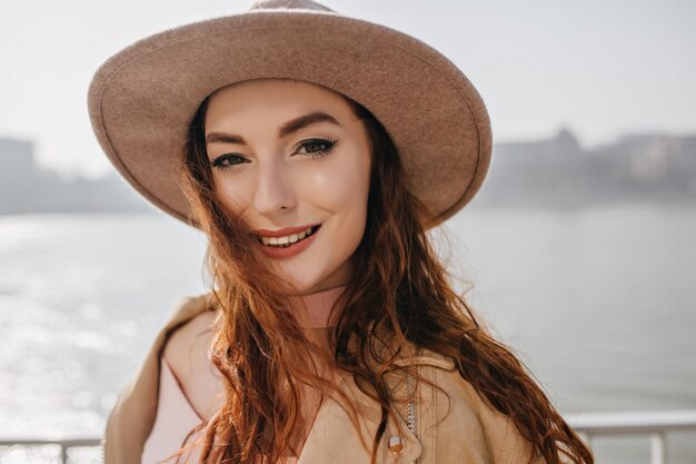 Close-up portrait of pleased ginger girl with white skin. Outdoor shot of cheerful caucasian lady with long hairstyle smiling on sea background in morning.