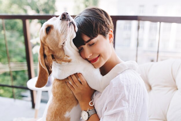 Close-up portrait of pleased woman with short brown hair embracing funny beagle dog with eyes closed