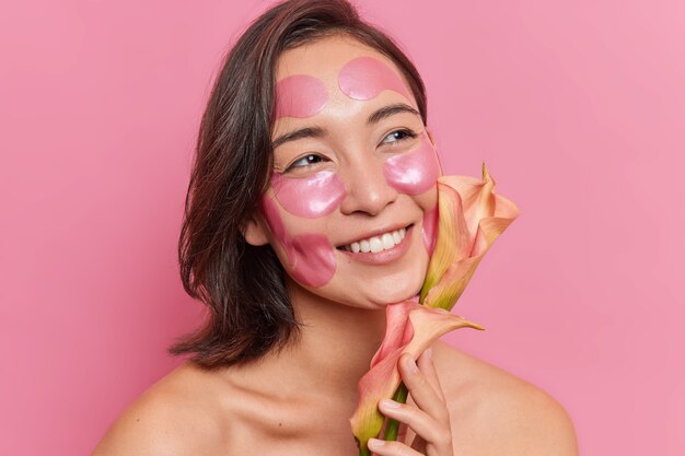 Close up portrait of pleased young Asian woman with toothy smile glad to get flower applies hydrogel patches on face to refresh skin stands shirtless against pink wall