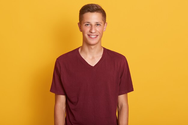 Close up portrait of pleased young guy dressed maroon casual t shirt, looking smiling at camera, expresses happyness, model posing isolated on yellow. People, youth and lifestyle concept.