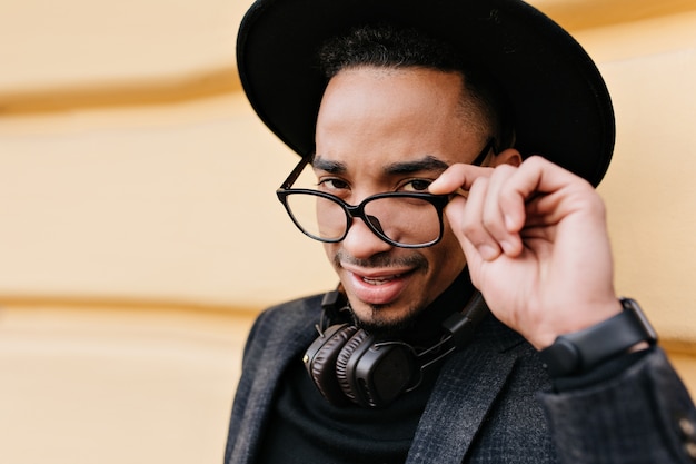 Ritratto del primo piano dell'uomo africano positivo che tocca scherzosamente i suoi occhiali. colpo esterno dell'elegante modello maschio nero in abbigliamento alla moda isolato sulla strada della città.