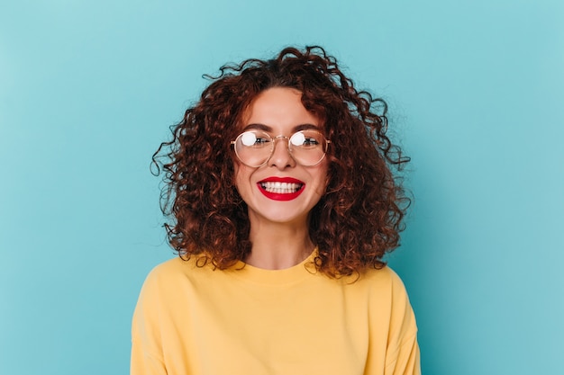 Close-up portrait of positive, dark-haired, curly girl in glasses. Woman with red lipstick dressed in yellow sweatshirt laughs wholeheartedly against blue space.