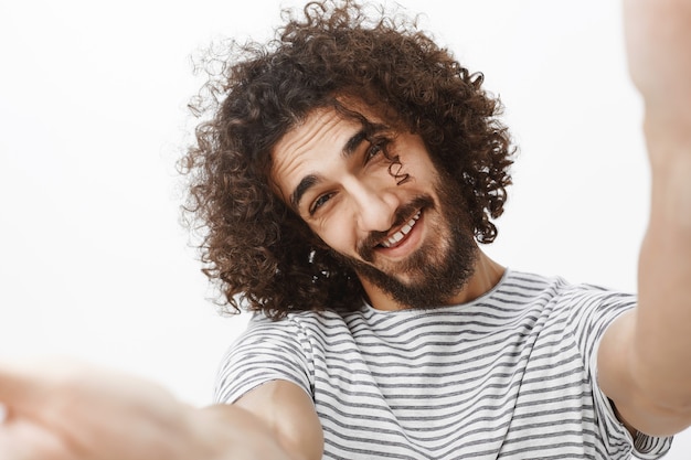 Close-up portrait of positive handsome emotive guy with beard and stylish curly haircut, pulling hands towards