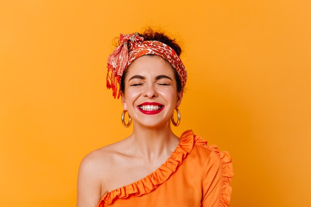 Close-up portrait of positive woman with red lips dressed in blouse with bare shoulder and headband laughing with closed eyes on isolated space.