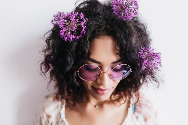 Close-up portrait of pretty african woman in sunglasses posing with eyes closed. wonderful female model with dark hair isolated with purple flowers.