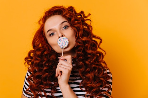 Ritratto del primo piano della ragazza graziosa dello zenzero con gli occhi azzurri che posano con il grande lecca-lecca sullo spazio arancione.