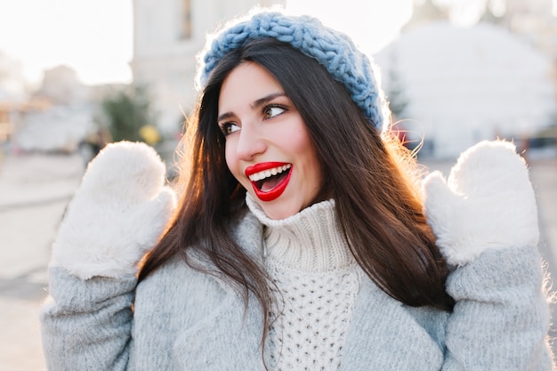 Close-up portrait of pretty girl with black long hair posing with hands up in cold day. Outdoor photo of cute european lady in blue hat and white gloves enjoying winter weekend.