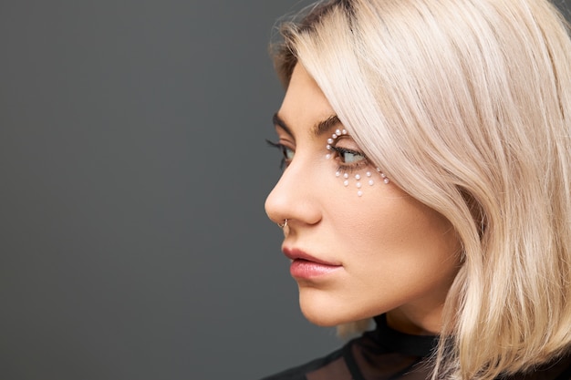 Close up portrait of pretty girl with blonde hair, facial piercing and artistic make up having thoughtful pensive look, posing against blank wall  with copy space for your text
