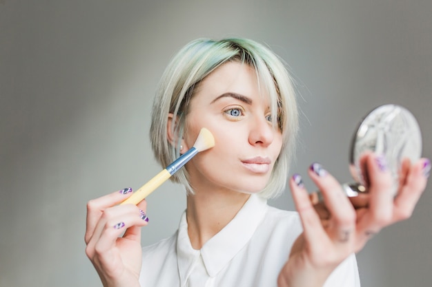 Close-up portrait of pretty girl with short white hair on gray background. She wears white dress, looks to the mirror in hand and powders face.