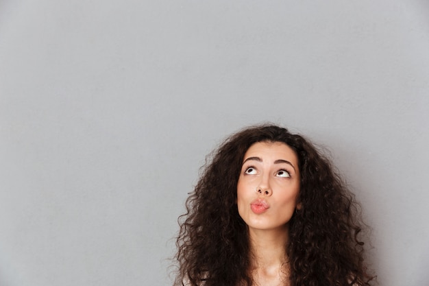 Free photo close up portrait of pretty lady having cool curly hairstyle looking up with twisted lips over grey wall