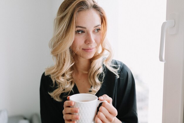 Close up portrait of pretty lovely girl with a cup of tea in the morning at home. She is sitting on the window and dreamy looking away