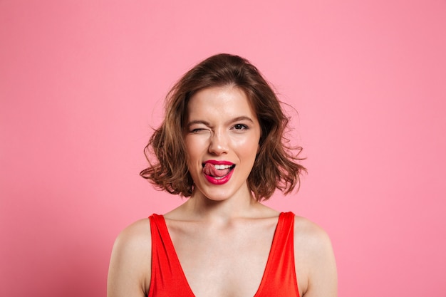 Close-up portrait of pretty playful woman winks one eye, showing tongue, , isolated on pink 