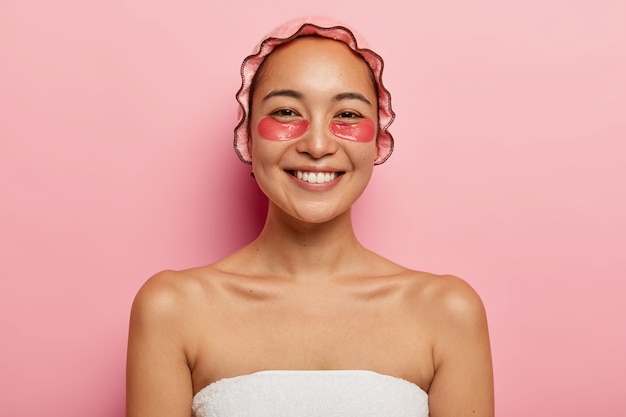 예쁜 웃는 여자의 초상화를 닫습니다 미용 치료, 수건에 싸서 분홍색 샤워 캡을 착용하고 주름을 줄이기 위해 눈 밑에 콜라겐 패드가 있으며 실내에 서 있습니다. 뷰티 개념.