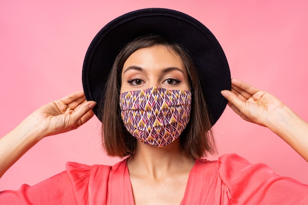 Free photo close up portrait of  pretty woman dressed protective stylish face mask. wearing black hat and sunglasses. posing over pink wall