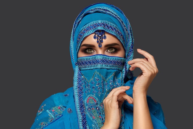 Close-up portrait of a pretty woman with beautiful smoky eyes wearing a blue hijab decorated with sequins and jewelry. She is gesticulating and looking at the camera on a dark background. Human emotio