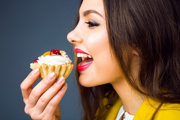 Close up portrait of pretty young brunette woman with bright makeup eating tasty cake with berries and cream.