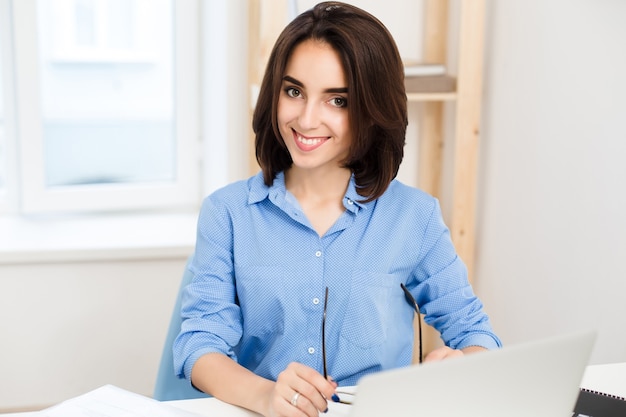 Close-up portrait of a pretty young girl in a blue shirt. She is sitting at the table in office and smiling to the camera.