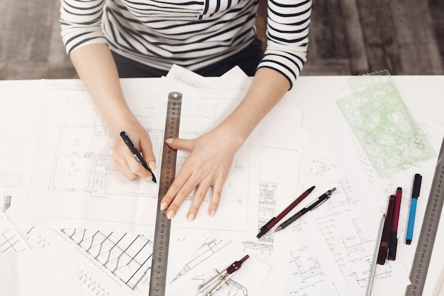 Free photo close up portrait of professional young beautiful female architect in striped clothes, doing her drawings with ruler and pen, working with interest on new project.