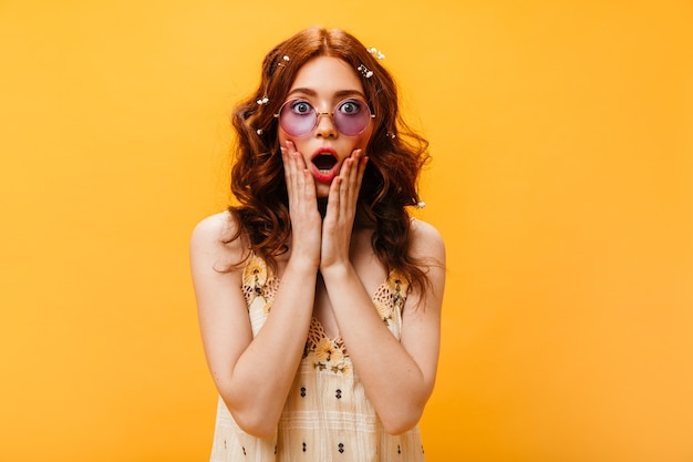 Close-up portrait of red-haired woman with wild flowers in her hair looking surprised into camera. Woman in lilac glasses posing on isolated background.