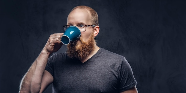 Foto gratuita ritratto ravvicinato di un maschio barbuto rosso beve caffè con gli occhiali vestito con una t-shirt grigia, isolato su sfondo scuro testurizzato.