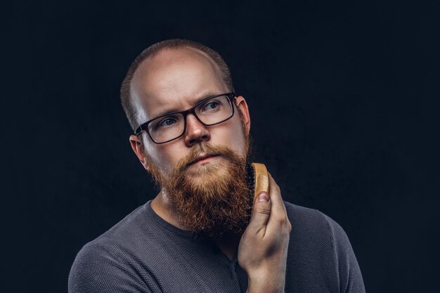 Крупным планом портрет рыжеволосого бородатого мужчины в очках, одетого в серую футболку, заботящегося о своей бороде с помощью щетки для бороды. Изолированные на темном текстурированном фоне.