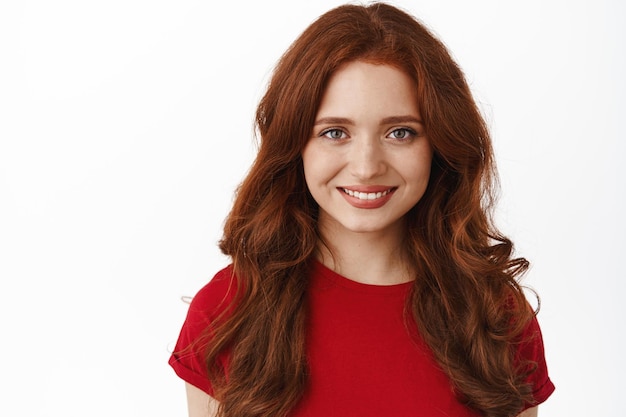 Close up portrait of redhead curly woman, smiling with white teeth perfect smile, looking confident and hopeful, standing in red t-shirt against white background.