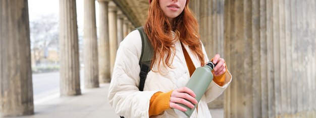 Close up portrait of redhead girl tourist stands on street opens thermos with hot drink rests and