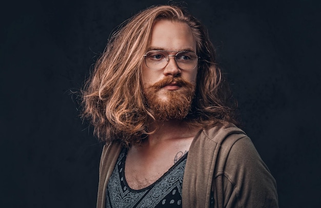 Close-up portrait of a redhead hipster male with long luxuriant hair and full beard dressed in casual clothes standing in a studio, looking away. isolated on a dark background.