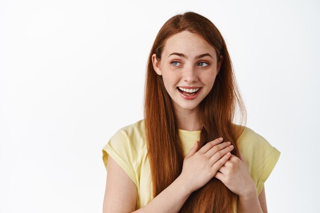 Close up portrait of redhead woman holding hands on heart, looking left dreamy, thinking of something romantic or love, standing over white background.