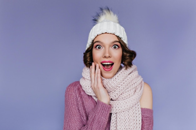 Ritratto del primo piano della donna riccia raffinata in cappello bianco. ragazza europea estatica con i bei occhi che posano in sciarpa sveglia.