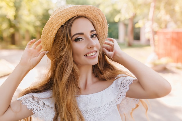 유행 여름 모자 웃 고있는 세련 된 숙 녀의 클로즈업 초상화. 우아한 반지와 흰색 옷을 입고 꽤 긴 머리 소녀의 야외 사진.
