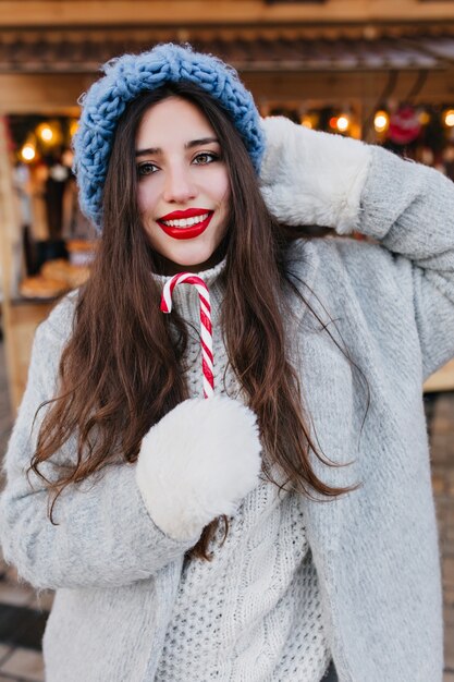 Крупным планом портрет романтичной европейской девушки с темными волосами, позирующей со сладким рождественским леденцом. Фото красивой кавказской девушки-модели в белых перчатках и синей шляпе, развлекающейся