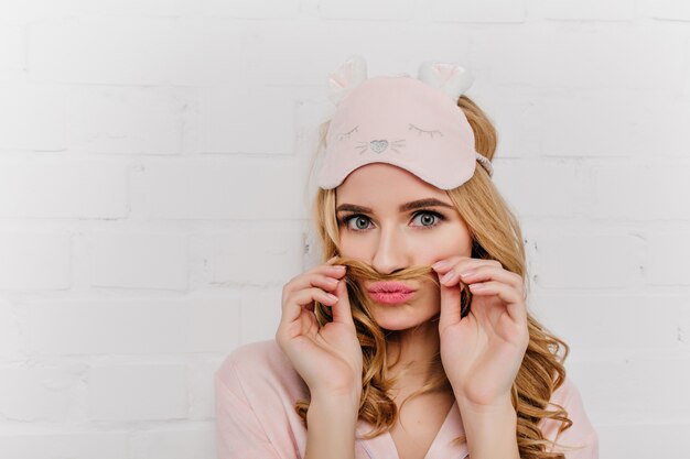 Close-up portrait of romantic pale girl in eyemask. Positive fair-haired woman wears pink pajama posing on light wall.