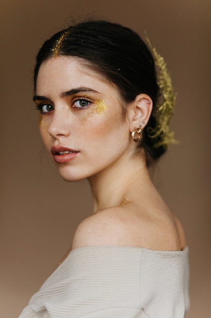 Close-up portrait of romantic young woman. Debonair pretty girl with golden accessories posing on dark wall.