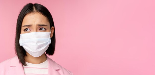 Close up portrait of sad frowning asian businesswoman in medical face mask looking left at copy spac