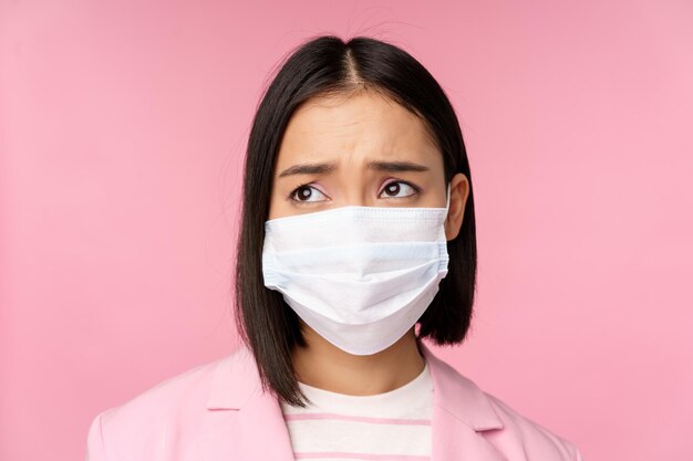 의료용 얼굴 마스크를 쓴 슬픈 아시아 여성 사업가의 초상화를 닫고 분홍색 배경 위에 서 있는 실망스러운 감정으로 카피 공간을 왼쪽으로 바라보고 있습니다.