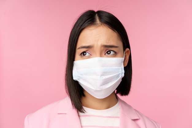Close up portrait of sad frowning asian businesswoman in medical face mask looking left at copy space with disappointed upset emotion standing over pink background