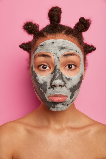 Free photo close up portrait of sad mixed race young woman looks at herself in mirror, applies clay mask on face, stands with bare shoulders, cares about body and complexion, isolated over pink wall.