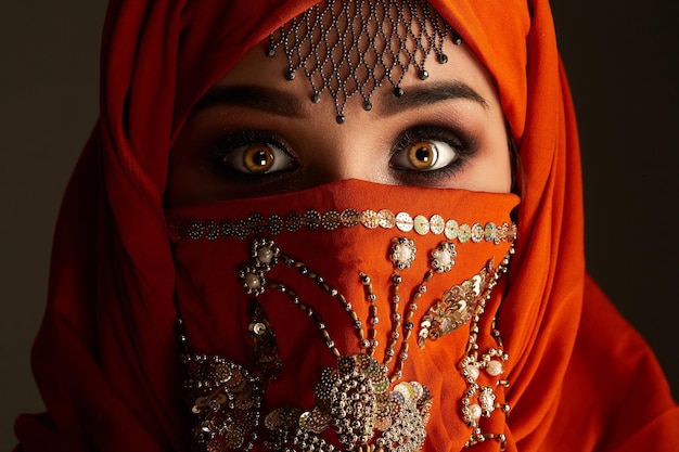 Ritratto ravvicinato di una giovane ragazza spaventata con occhi fumosi e gioielli sulla fronte, che indossa l'hijab di terracotta decorato con paillettes. sta guardando la telecamera su uno sfondo scuro. emozione umana