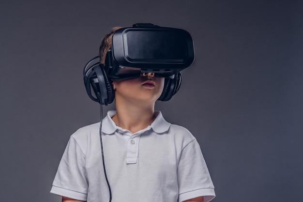 Close-up portrait of a schoolboy dressed in a white t-shirt standing in a virtual reality glasses at a studio. Isolated on a gray background.