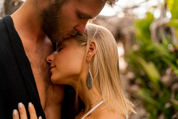 Close up portrait of seductive couple in tropical garden Beautifyl blond woman in hat and boho outfit hugging with her handsome boyfriend Summer romantic moments